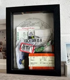Adventures Memory Box frame filled with travel keepsakes, including boarding passes, train tickets, currency and photos, displayed against a world map background with ‘Each day is a one ticket adventure’ printed on the glass.