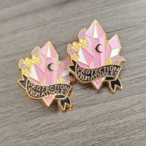 Two enamel pins featuring pink and white crystal clusters with a black crescent moon, surrounded by small yellow and purple flowers. A black ribbon banner with gold trim reads “Protection From Assholes.” The pins rest on a wooden surface.