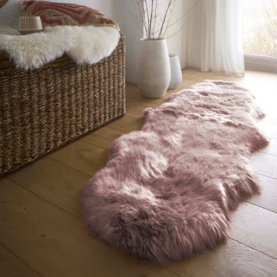 A Genuine Sheepskin Rug in the Adobe Rose color draped over a woven bench, styled with a pink cushion, ceramic mug, and soft natural décor, with a long pink faux‑fur rug on the floor in a warm, light‑filled room.