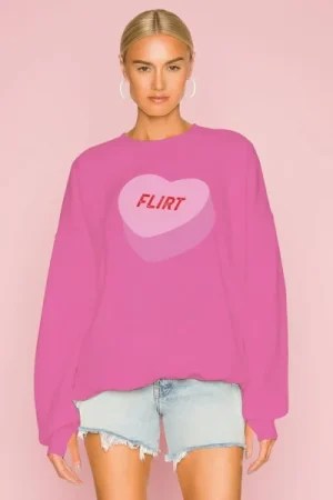 A model wears The Laundry Room Sweethearts Flirt Sweater, an oversized bright‑pink pullover featuring a light‑pink candy‑heart graphic with the word “FLIRT” in bold red lettering. Styled with light‑wash frayed denim shorts and white hoop earrings, the look is set against a soft pink background for a playful, romantic aesthetic.