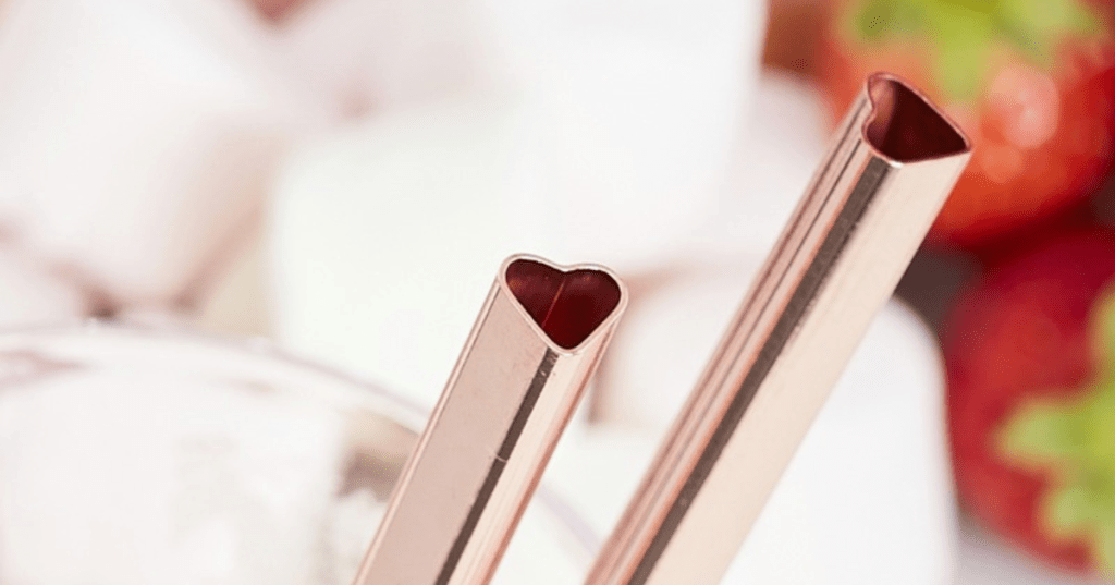 Two rose gold metal straws with heart-shaped openings rest inside a clear glass, set against a soft-focus background of strawberries and marshmallows. The romantic design and warm metallic finish make these reusable straws ideal for Valentine’s Day, weddings, or festive gifting.