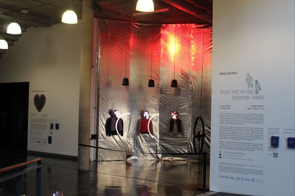 Gallery installation for Nina Davies’ Meet Me in the Digital Twin, featuring three illuminated mannequin torsos against a reflective metallic backdrop with red lighting, pendant lamps overhead, and exhibition text displayed on the adjacent wall.