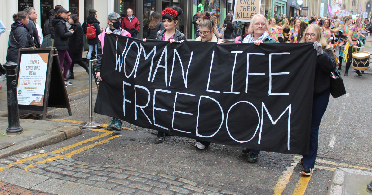 Marchers walk down Liverpool’s Bold Street during a Women’s March, carrying a large black banner reading “Woman Life Freedom” at the front of the procession. Behind them, protesters hold additional signs as drummers and brightly dressed participants fill the street, creating a powerful scene of solidarity and activism.
