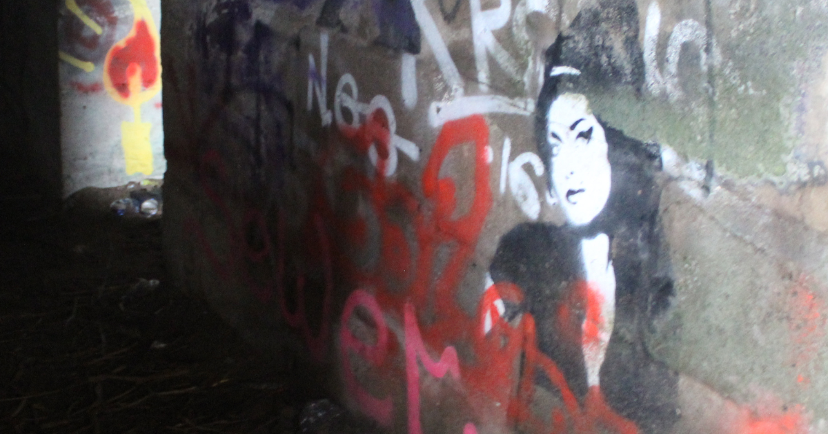 Graffiti artwork inside the Fort Crosby bunker showing a black‑and‑white stencil‑style portrait resembling Amy Winehouse on a concrete wall, surrounded by colourful spray‑paint tags and debris on the ground in the dim, abandoned space.