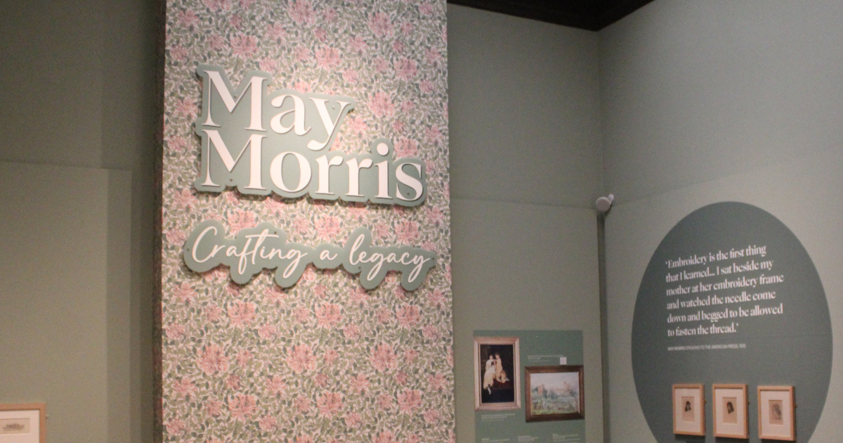 Exhibition display featuring the title ‘May Morris: Crafting a Legacy’ on a floral-patterned wall, with a circular section beside it showing a printed May Morris quote and framed artworks below.