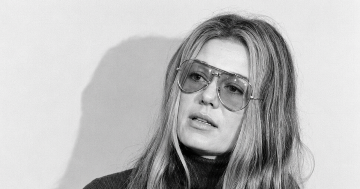 Black‑and‑white portrait of Gloria Steinem wearing large aviator‑style glasses and a dark high‑neck top. Her long straight hair falls past her shoulders as she looks thoughtfully to the side, with soft lighting casting a subtle shadow on the wall behind her.