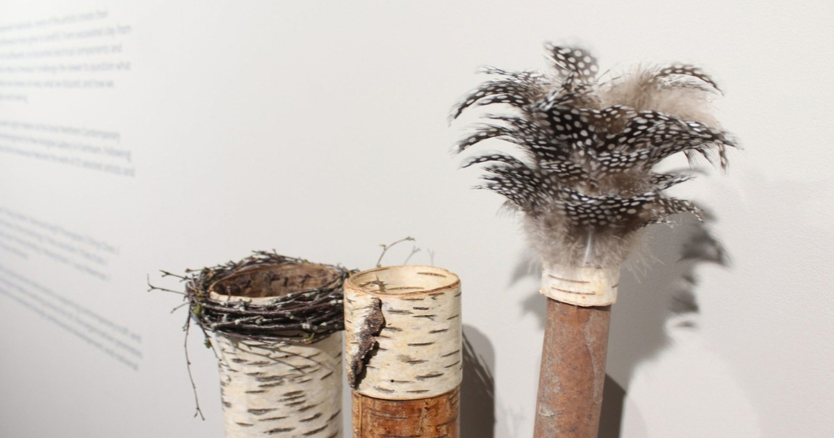 Jane Bevan collecting pots, including two birch‑bark cylinders—one wrapped with a delicate nest of twigs—and a taller rust‑toned stick topped with black‑and‑white spotted feathers. Their foraged textures and woodland assemblage reflect Bevan’s signature practice of crafting intimate pots from bark, twigs, and found organic elements.
